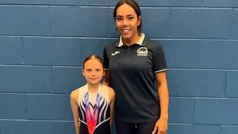 Ana Metcan Mikayla Metcan standing with her coach. She is wearing a gymnastics leotard and has a medal around her neck. Her coach is wearing a black polo shirt.