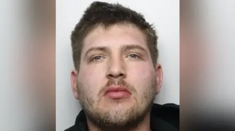 A headshot of a young man with short brown hair, a short beard and moustache and grey eyes.