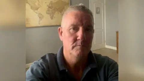 Mark Hemphill A selfie of Mark Hemphill, he is indoors wearing a navy polo shirt. A world map is on the wall behind him.