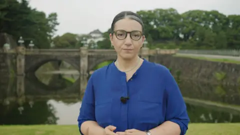 BBC Japan correspondent Shaimaa Khalil in front of the Japanese Imperial Palace. She is wearing her hair back, glasses and a blue shirt.