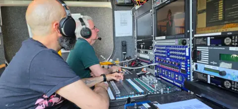 Two BBC engineers in the foreground helping to output Monday's edition of Look East, with various dials and screens in front of them on the desk