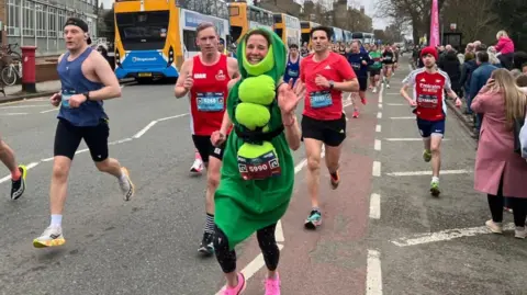 Cultivating Change A woman dressed as a pea running up a road in central Cambridge. There are three male runners immediately behind her and crowds of other runners further down the road. On the right, people have line the pavement to cheer the runners on. 
