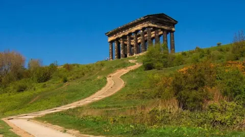fullframejpg A steep grassy hill with a path leading up to it. Planks have been used to create a series of steps along the path. There are people walking towards the top where there is a large dark stone monument with pillars, reminiscent of the Acropolis. The sky is bright blue with no clouds.