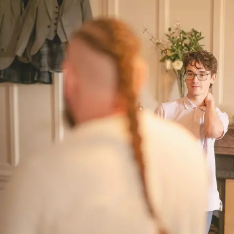 Duncan Ireland Photography A candid wedding photo. A young man is standing in his shirt with one arm around his own neck. In the foreground is the out-of-focus image of the rear of a large man with a pony tail. 