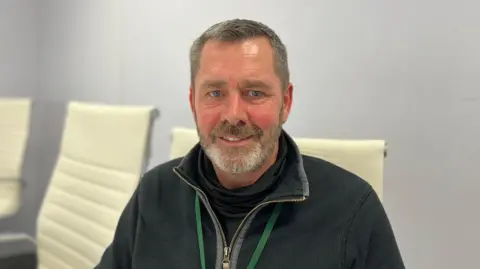 Deputy Marc Leadbeater - A man with black and grey hair looking at the camera. He's wearing a black quarter zip and a green lanyard. 