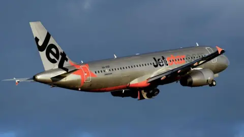 A silver plane with Jetstar's orange and black livery flies against a dark cloudy sky.