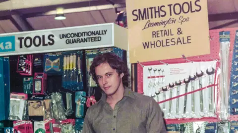 Laser Tools A man, Martin Smith, has short wavy brown hair and is wearing a brown shirt. He is standing in front of a stall with numerous tools around him. 