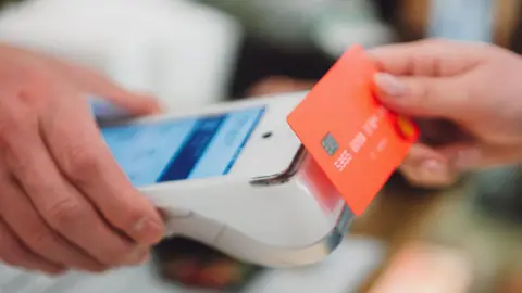 Getty Images Someone is holding out a card machine, someone else is holding a card towards it and making a contactless transaction
