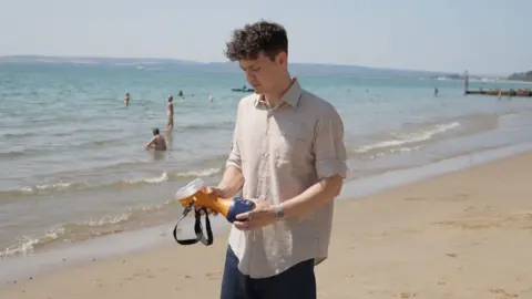 Rupert Murphy standing in front of the sea at Alum Chine in Bournemouth. He is wearing a white shirt which has multicoloured lines on it. He is stood on the sand. He is holding the Re-Warm device. It is orange with a blue base and black strap.