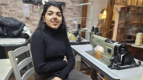 A woman sits in front of a sewing machine. She has long dark hair and is wearing a black jumper and dark grey trousers. Behind her a number of spools of thread can be seen.