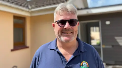 A man with grey hair and a beard, wearing sunglasses and a blue polo shirt with the Les Amis logo