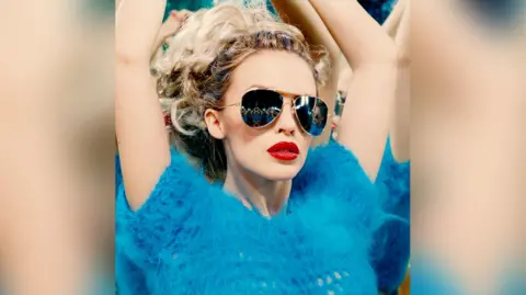 Norbert Schoerner Kylie Minogue with her arms in the air, wearing sunglasses and a fluffy blue jumper 