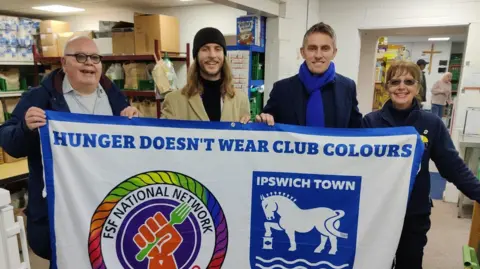 Ipswich Town Fans Supporting Foodbanks stand behind a large flag that they are holding. Ipswich Town manager Kieran McKenna stands with them holding the flag. They all wear winter coats and clothing. The white flag reads: Hunger doesn't wear club colours. It has the Ipswich Town logo on it.