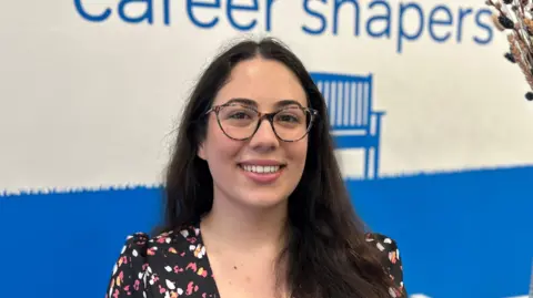 Veronica Da Costa with long brown hair and glasses. She's wearing a dress with a pink and white design and is standing next to a vase of flowers in a blue and white room. 