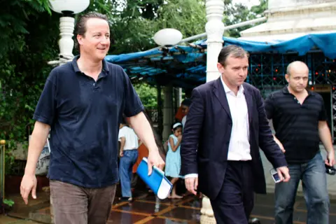 Press Association David Cameron, left, then the leader of the Conservative Party, and Steve Hilton, on the right, visit India in 2006. Cameron wears a navy blue polo shirt and brown pants, and Hilton wears a blue striped polo shirt. A man is also seen walking between them.