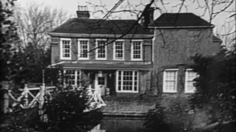 A black and white photo of the front of a house with a mill stream in front of it