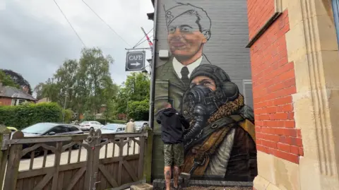 A graffiti mural on the side of a grey building. The artwork includes an airman wearing breathing apparatus and headphones. Above him is a spray painted man in uniform which is half completed. 
