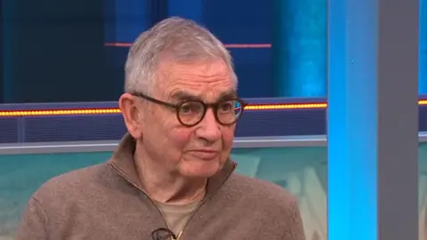 Older man with short grey hair and round glasses wearing a brown zip-neck jumper, speaking in a television studio with blue and orange lighting in the background