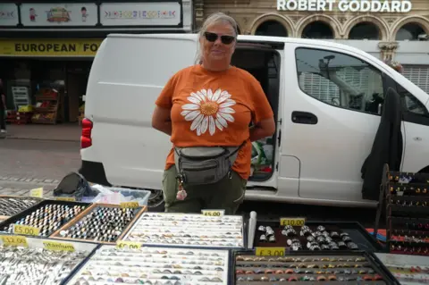 A woman with brown hair, tied back, is wearing sunglasses and an orange T-shirt with large daisy design as she stands behind a jewellery stall with a white van parked behind her.