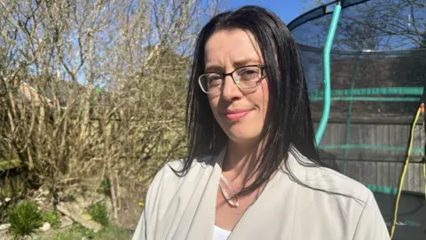 Chanel Walsh in the garden of her home. There is a trampoline behind her. She is smiling and looking at the camera. She has long dark hair past her shoulders and wear half-rimmed glasses.