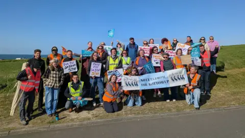 James Armstrong A group of teachers and support staff stand by the side of the road with banners and placards.