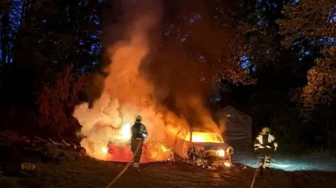 Firefighters tackling a large fire with big orange flames in the dark. There are two firefighters with hoses pointing towards the fire.