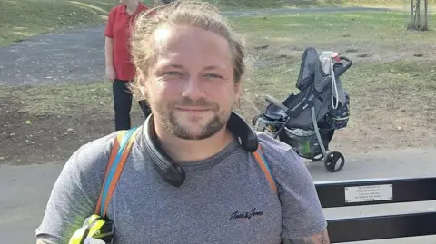 Ashley Southall Ashley Southall sat on a bench ready for his walk. He has a sports top on and a fan around his neck. There is a pushchair behind him in the background. He is smiling and has long hair and a goatee-style beard.