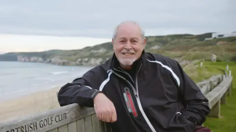 A man with short white hair smiles at the camera. He is wearing a black jacket and sitting on a bench outside. Behind him is a beach.