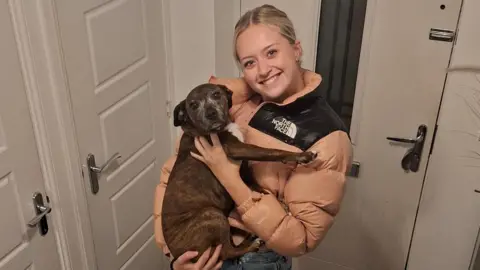 Helen and Lee Draper A young woman is holding a dog while smiling at the camera in a house, infront of the front door.