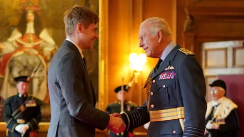 PA Media Swimmer Stephen Clegg wearing a grey suit shakes King Charles' hand. He is wearing a black jacket with a pale-coloured shirt and gold belt.