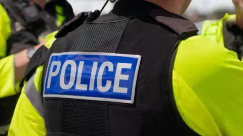 A police officer with their back turned. Image shows yellow high vis jacket and black vest with 'Police' written on.