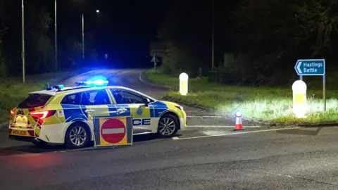Dan Jessup A police car at the scene of a road traffic accident in Robertsbridge, East Sussex. The car is parked across the road, blocking access. The blue light is on.