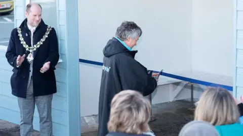 A woman in a dry robe cuts a blue ribbon with scissors that is across the entrance to a blue and white beach hut. A man in a black coat, grey trousers and mayoral chains claps nearby.