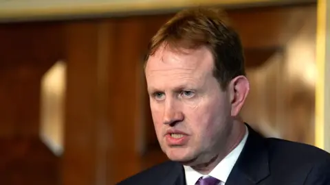 PA Media Jim O'Callaghan is wearing a dark suit, white shirt and pink tie. He has dark short hair and is clean shaven. He is speaking with a serious expression, looking to the right of the camera.