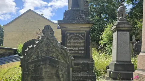 Hayley Coyle/BBC Historic gravestones and memorial monuments in a cemetery, surrounded by tall grass and mature trees on a bright day.