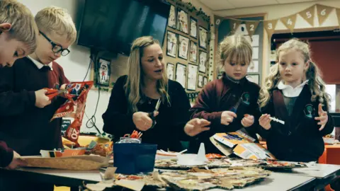 Two boys and two girls are standing next to a table as they cut and glue items of litter including cardboard boxes. They are wearing maroon school jumpers or cardigans. The boy second from the left has dark glasses. All four have blonde or brown hair. They are with a female member of staff who is also holding a pair of scissors. She has blonde hair and is wearing a back top. A television is mounted to a wall behind them. The space is also decorated with a number of drawings.