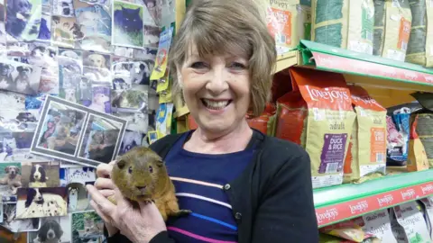 Barbara Mullaney, who has short brown hair and is wearing a blue and black outfit. She has a big smile and is stnaidng in a pet shop holding a guinea pig.