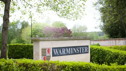 Warminster School A sign on a thick brick rectangular object which says 'Warminster' with a hedge and trees in the background.