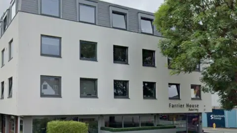 A rectangle-shaped building which is light grey and over several floors. There are green bushes outside and a sign on the building says Farrier House. 