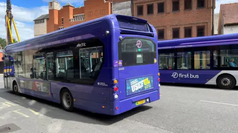 This image shows purple-painted buses heading in both directions along York's Rougier Street, where there are plans to restrict access to some vehicles to speed up bus journey times. A crane can be seen to the left of the picture.