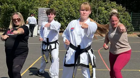 Mixture of people in taekwondo uniform (dobok). Some are wearing black belts and some people are wearing t-shirts and gym wear and putting their left fist out. 