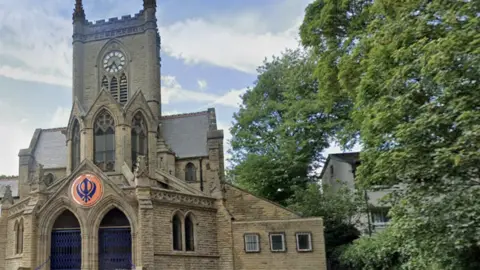 A brown stone church building with a clock tower, and a large orange and blue Sikh symbol over the front door.