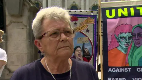 A woman with short grey hair, pictured from the shoulders up. She is wearing a navy top, a necklace and glasses. There are anti-racism posters hung up behind her.