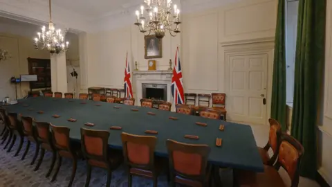A picture of the Cabinet Room in Downing Street. There is a large table in the centre of the room, surrounded by chairs. At the centre there is a mantelpiece with two union flags on either side. In the far corner, there is one bookcase. 