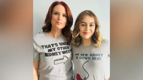 PA Media Emma Palmer is standing side-by-side indoors with Maya. Emma has red hair and is wearing a grey T-shirt with writing printed, that reads THAT'S WHERE MY OTHER KIDNEY WENT. It is accompanied with the drawing of an arrow, pointing to her left. On her left is Maya, who has blonde hair and is smiling. She has a grey T-shirt reading MY NEW KIDNEY IS DOWN HERE, with an arrow pointing to a drawing of a kidney, which is coloured in red.