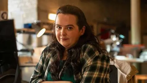 BBC Storrie in a scene from the programme Dinosaur looks directly at the camera. She is wearing a check shirt and a lanyard which says Glasgow. She is sitting in a deserted office with an anglepoise lamp seen behind her.