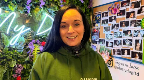 A woman smiling at a camera as she stands in a brightly decorated shop. She has black hair and is wearing a green hoodie. She is standing in front of a wall of fake plants, with a neon sign to her left. To her right are lots of Polaroid-style photos of dogs which are pinned up on the wall. 