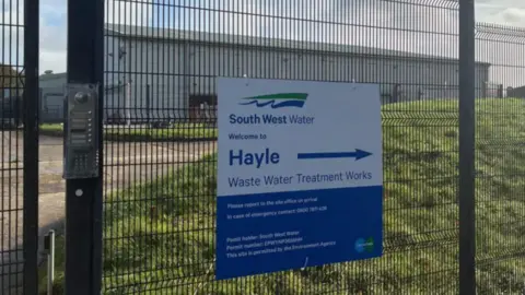 A mesh fence surrounds a warehouse type building. A sign on the fence says it's owned by 