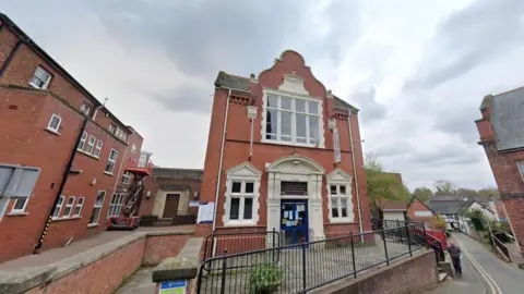 A Google street view image showing a two storey library building in the middle of a town, with a black fence in front of it. A narrow road is on the right with a man walking on the pavement.