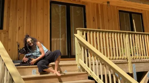 Erica Cirino Wearing sunglasses and holding her black dog, Erica Cirino sits on the wooden steps of her home. 